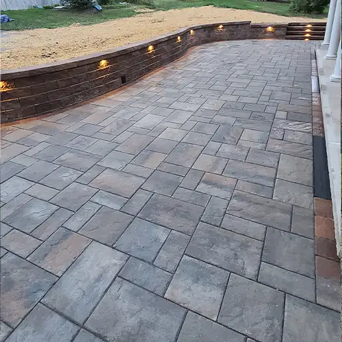 this is an image of a patio paved in brown paver stones with a curved retaining wall with built in lighting by cappellalandscaping.com